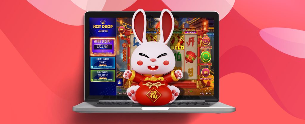 A 3D-animated cartoon rabbit wearing a red and gold Chinese emperor’s-style jacket is seen sitting on the keypad of an open silver laptop, with the screen behind showing the SlotsLV slots game, Year of the Rabbit Hot Drop Jackpots.